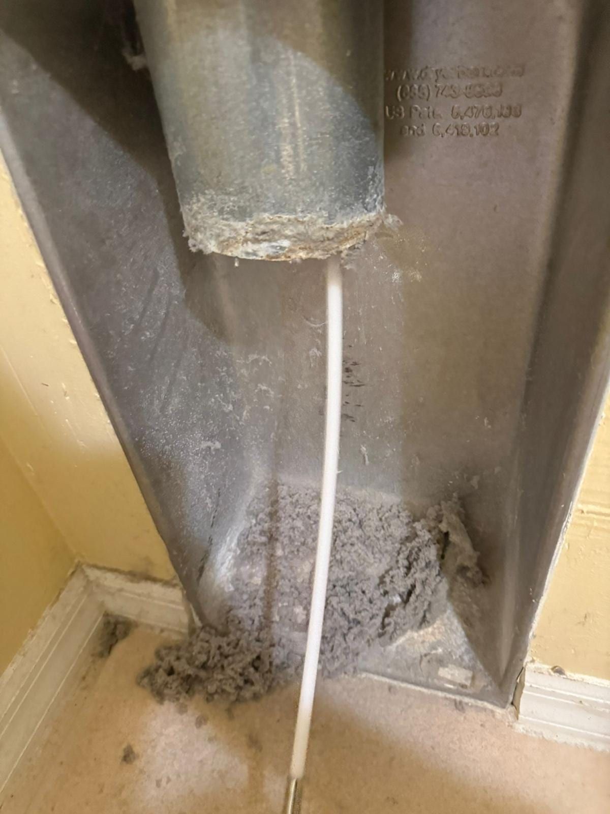 A dryer duct connector caked with lint at the collar, a clump of lint on the floor beside it and a rod pushed into the duct