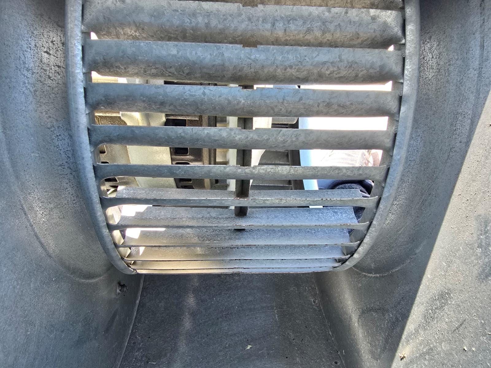 Looking along the inside of a galvanised duct trunk toward a slatted grille, daylight coming through the louvers